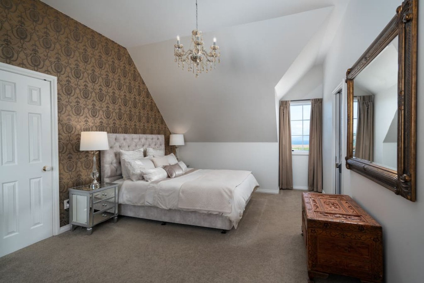 A bedroom with a large bed, white bedding, and multiple pillows. There are two bedside tables with lamps, a chandelier hanging from the ceiling, and a large ornate mirror on the wall. The room has a window with beige curtains and a view outside.