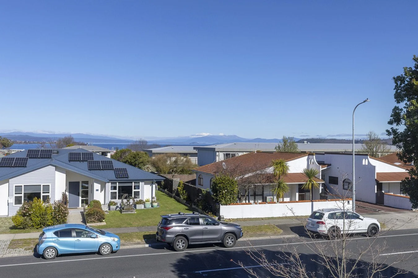 Residential neighborhood with two houses, a street with three parked cars, and distant mountains with snow caps under a clear blue sky.