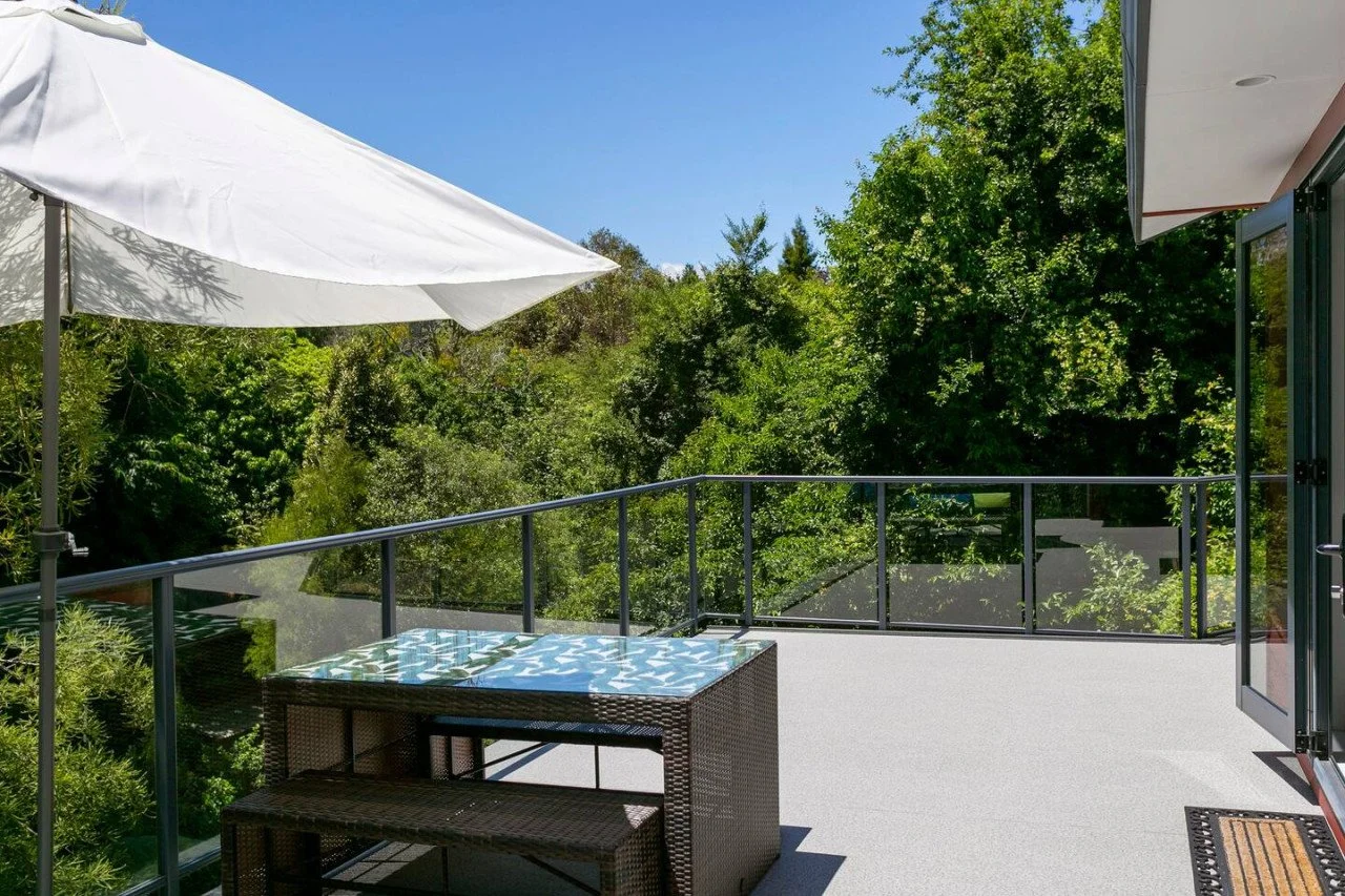 Balcony with a wicker table and bench, an open door leading outside, a large white umbrella, and green trees in the background under a blue sky.