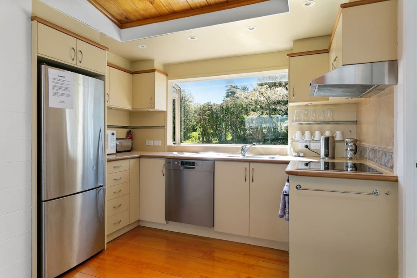 Kitchen with cream-colored cabinets, stainless steel refrigerator, dishwasher, and oven, open window revealing trees and greenery outside, with sunlight illuminating the space.
