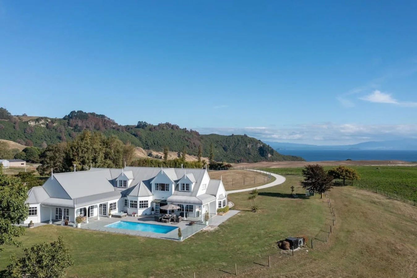 A large white house with multiple gabled roofs and a swimming pool in the backyard, surrounded by green lawns and trees, with hills and water in the distance under a clear blue sky.