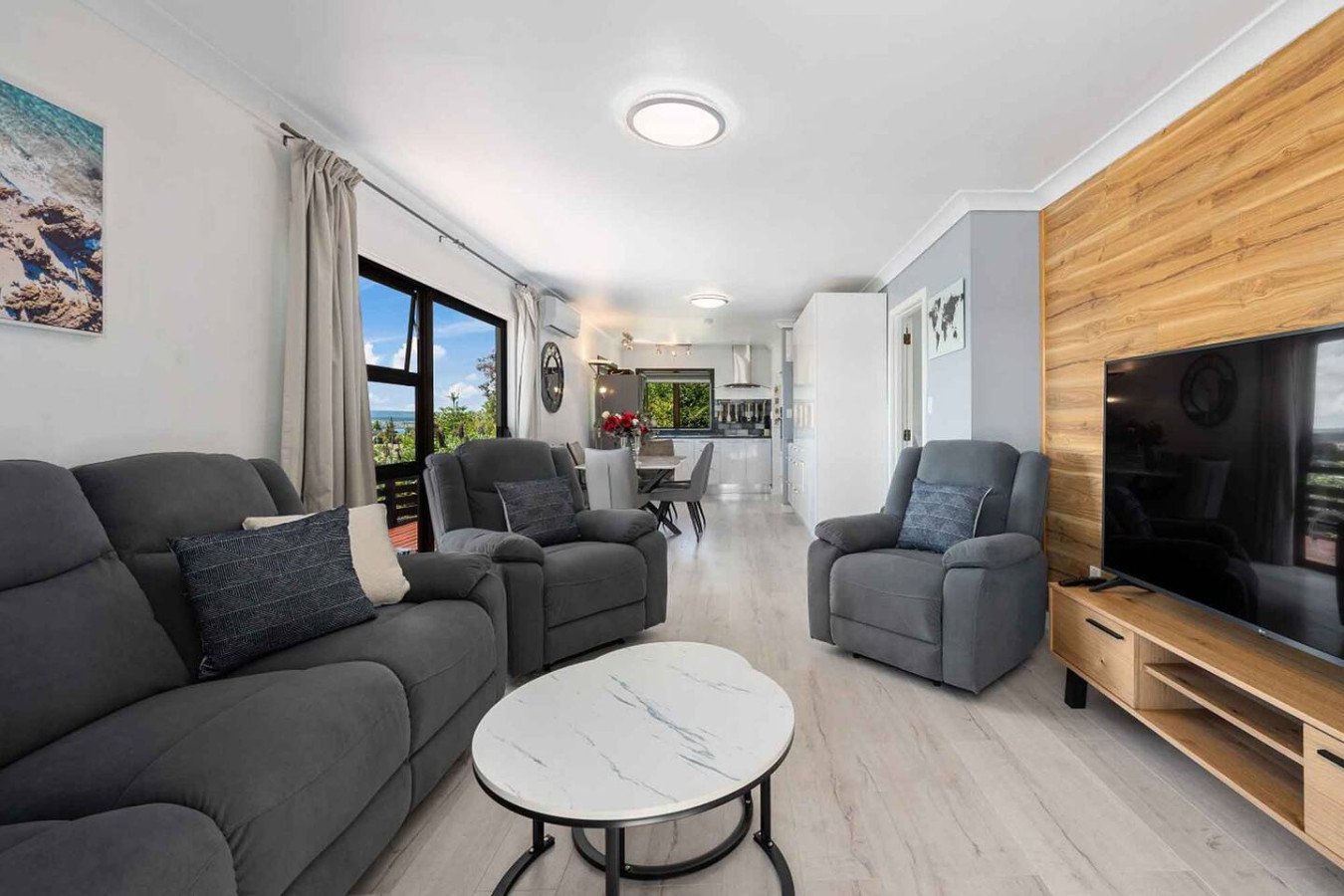 Living room with gray couches, a TV on a wooden stand, dining area, kitchen, window with view, and light-colored wooden floor.