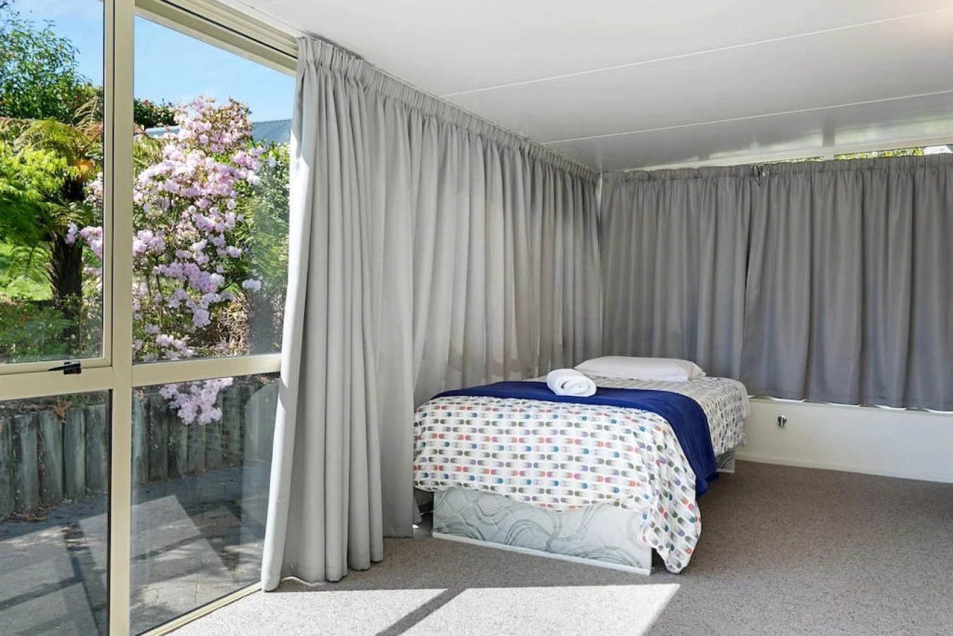 A bedroom with a large window covered partly by gray curtains, showing pink flowering trees outside. Inside, there is a bed with a colorful polka dot bedspread, white pillows, and a rolled white towel.