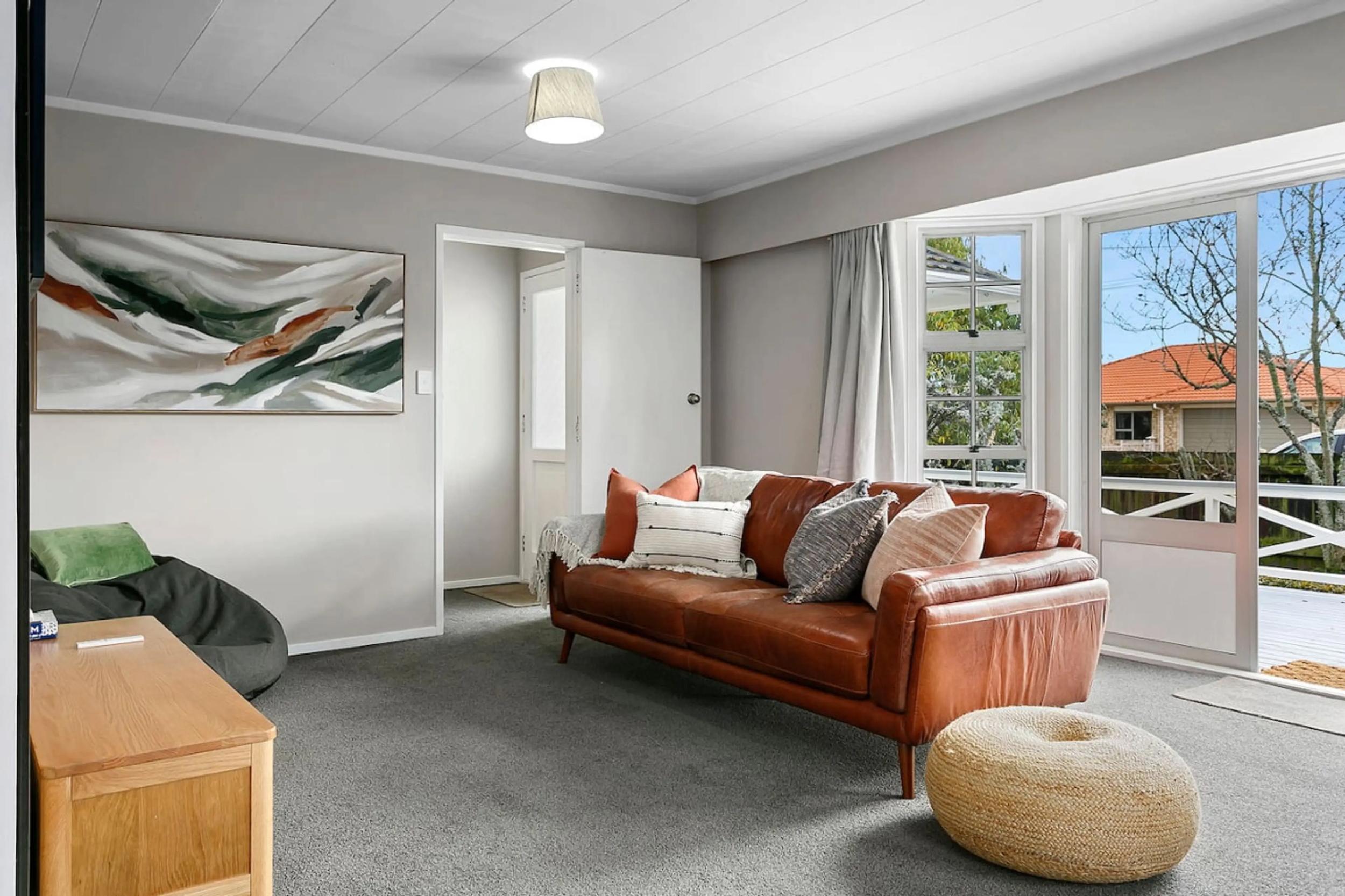 A living room with gray carpet, a white ceiling, and large windows showing an outdoor deck and trees. There is a brown leather sofa with multiple pillows, a small wooden table, a green bean bag chair, a woven round pouf, a wall-mounted abstract paint