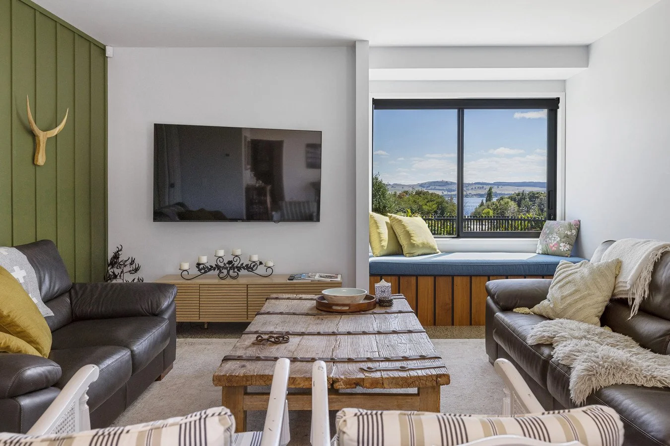 Living room with leather sofas, a wooden coffee table, a wall-mounted TV, and a window seat with a view of trees and water.
