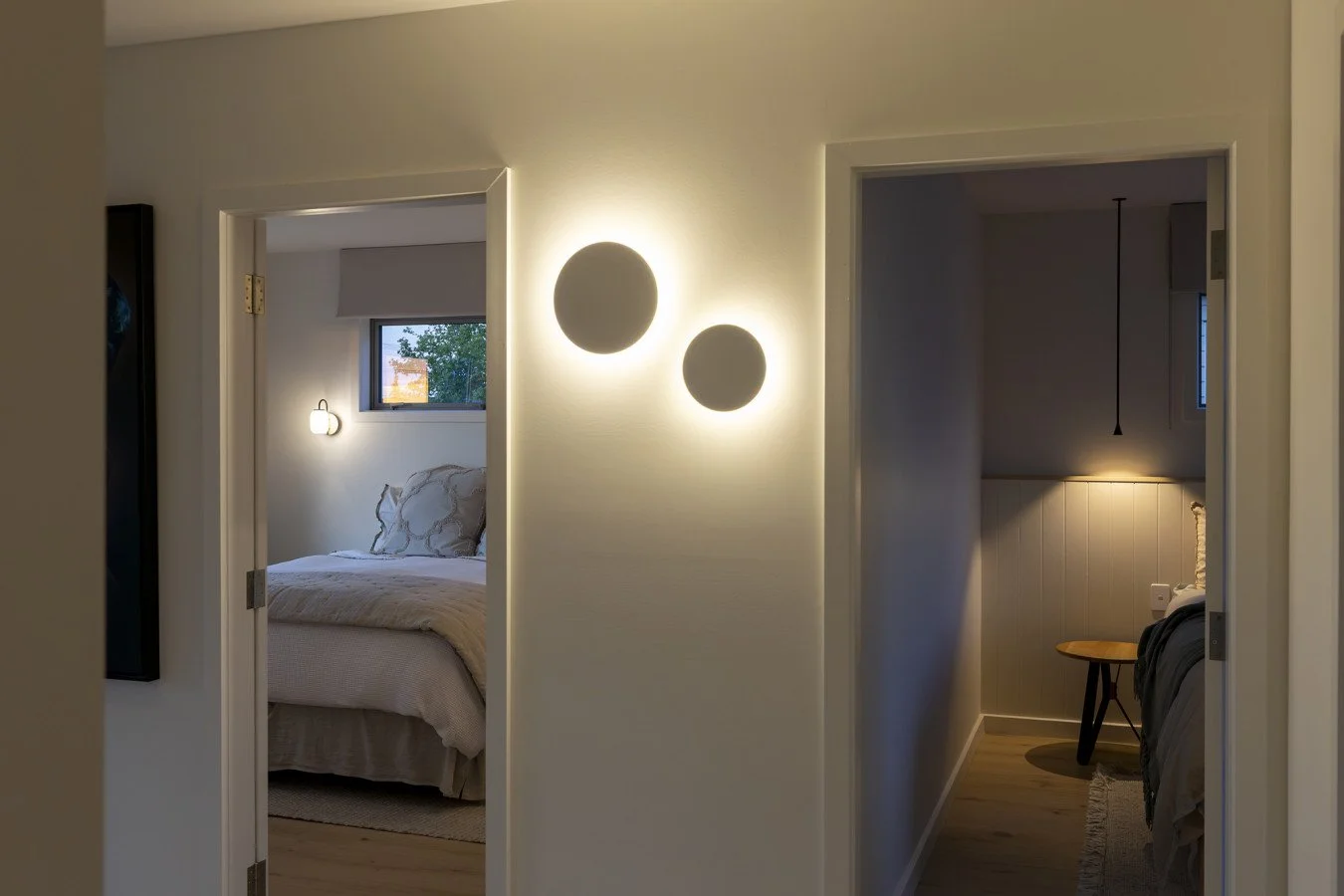 A hallway with two bedroom doorways. The wall between has two round wall lights, one larger and one smaller, and the bedroom on the left has a bed with pillows and a window. The bedroom on the right has a hanging pendant light and a small table.
