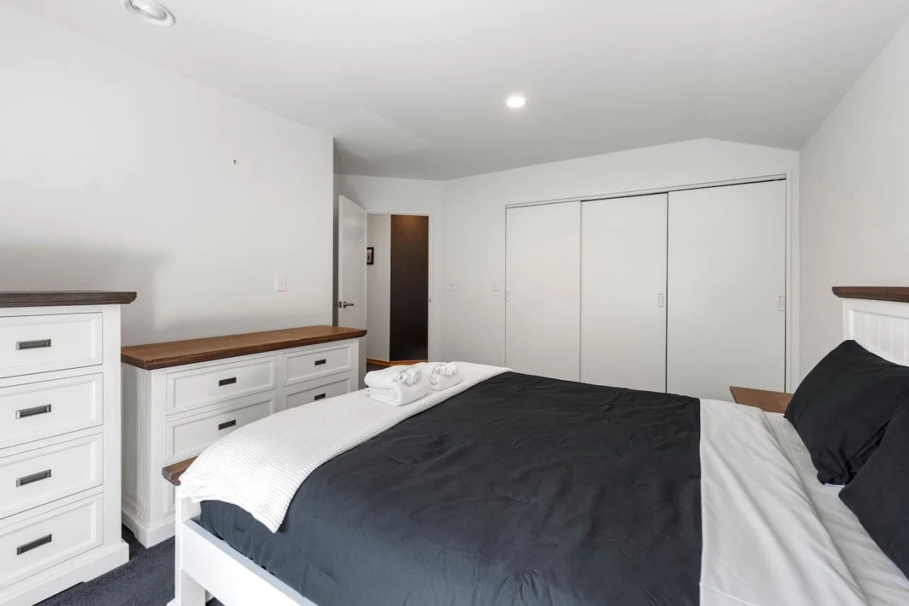 Empty bedroom with white walls, a bed with black and white bedding, white dresser and cabinets, and closed closet doors.
