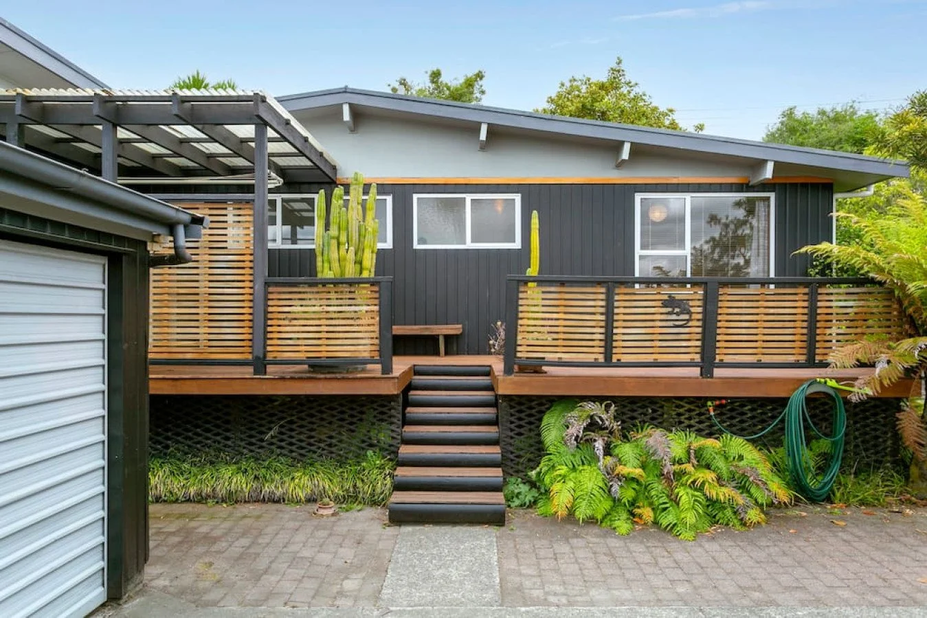 Modern black house with a wooden deck, stairs, and lush green plants, including cacti and ferns, in a suburban setting.