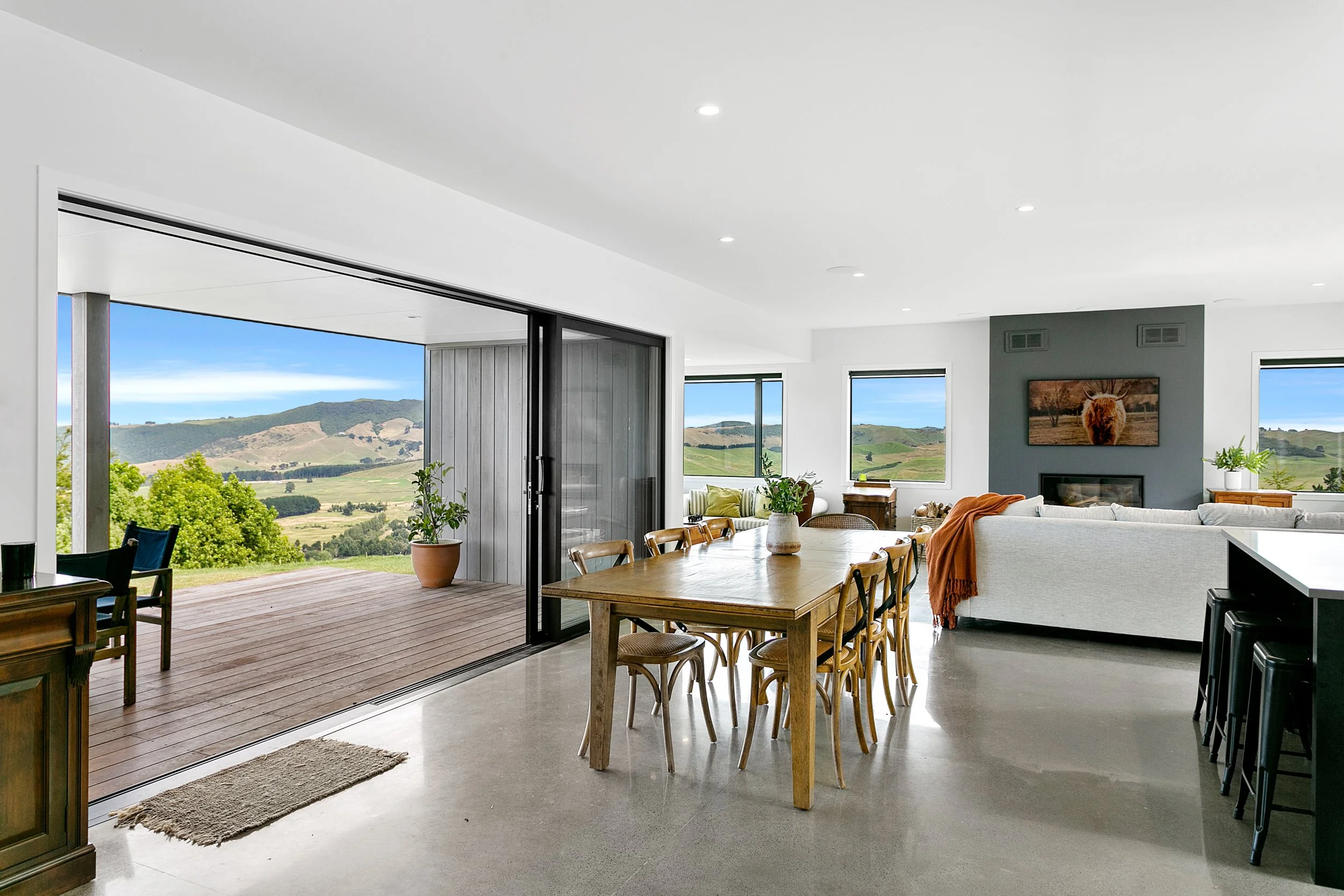 Bright open living and dining area with large sliding glass doors opening to a scenic outdoor deck with rolling hills view; modern furniture, neutral decor, and natural light.