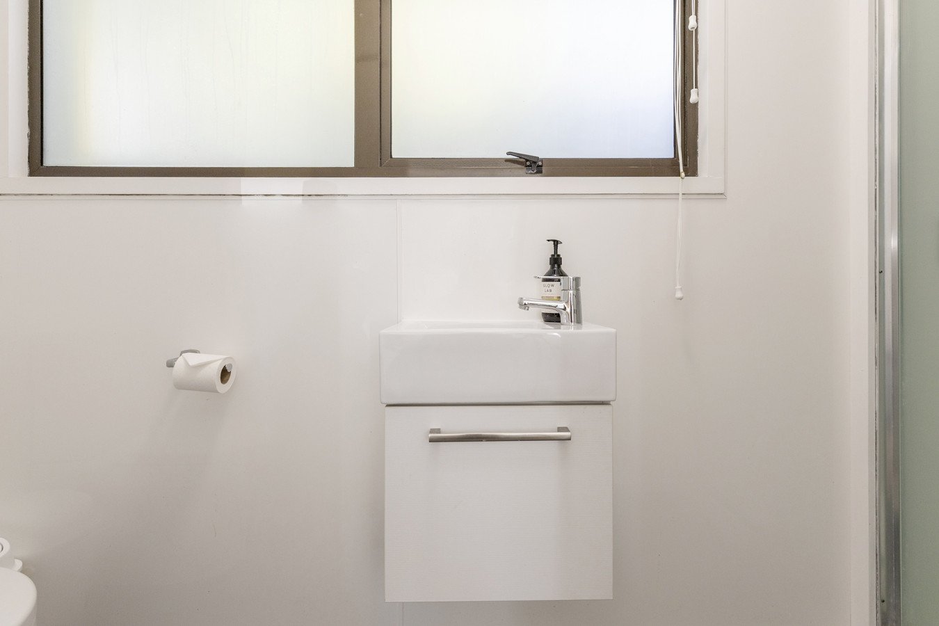 Small white bathroom sink with a cabinet underneath, a single-handle faucet, a frosted window above, and a toilet paper roll on the wall to the left.