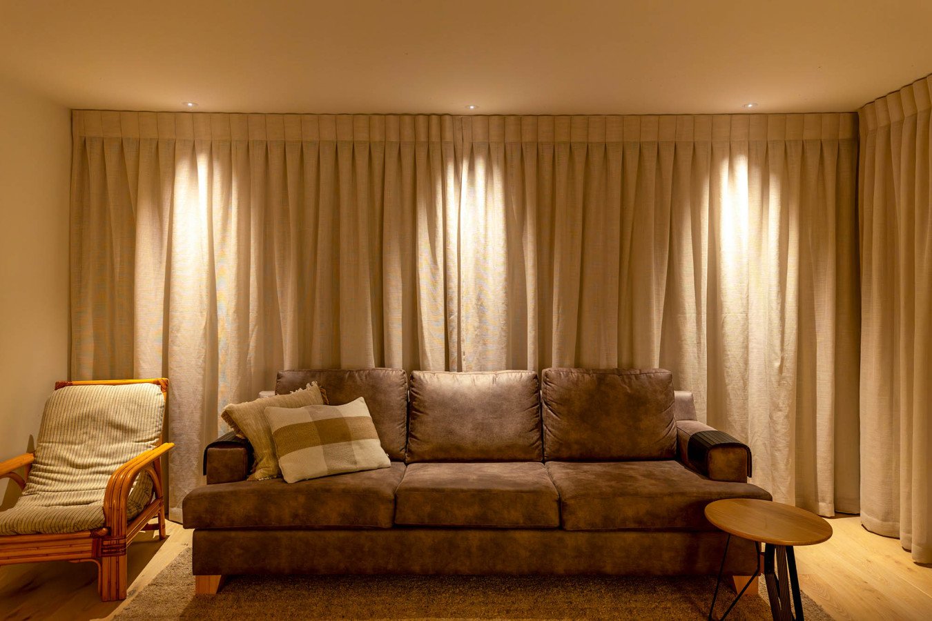 Living room with a brown sofa, beige curtains, a wooden side table, and a rattan armchair.