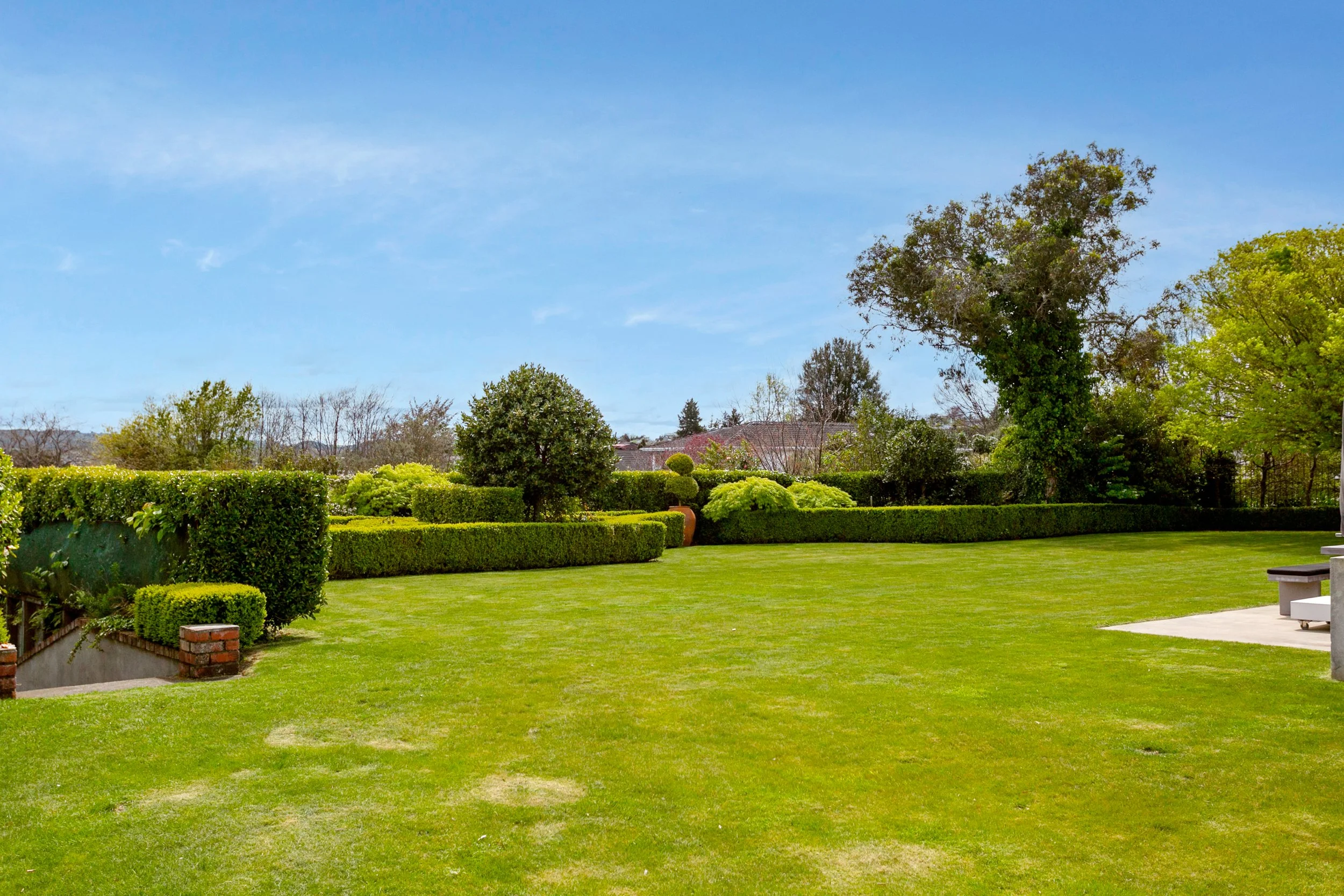 A landscaped garden with a large, well-maintained green lawn, surrounded by neatly trimmed bushes and trees, under a bright blue sky.