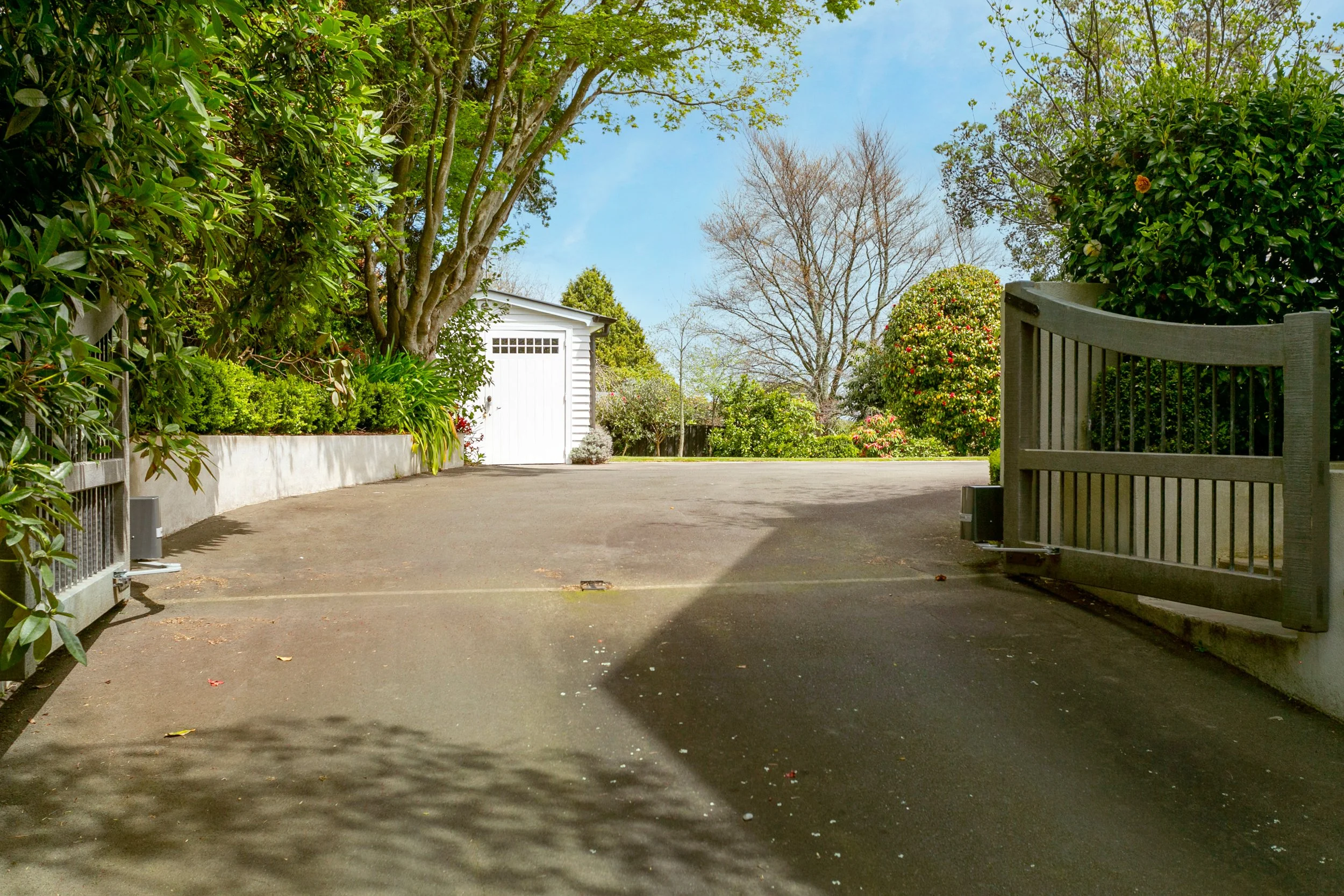 A driveway leading to a white garage with a handle, surrounded by green bushes, trees, and a clear blue sky.