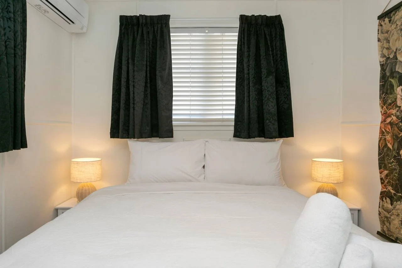 A cozy hotel bedroom with a neatly made bed, white bedding, and two matching lamps on bedside tables, with dark curtains and a window with blinds behind the bed.