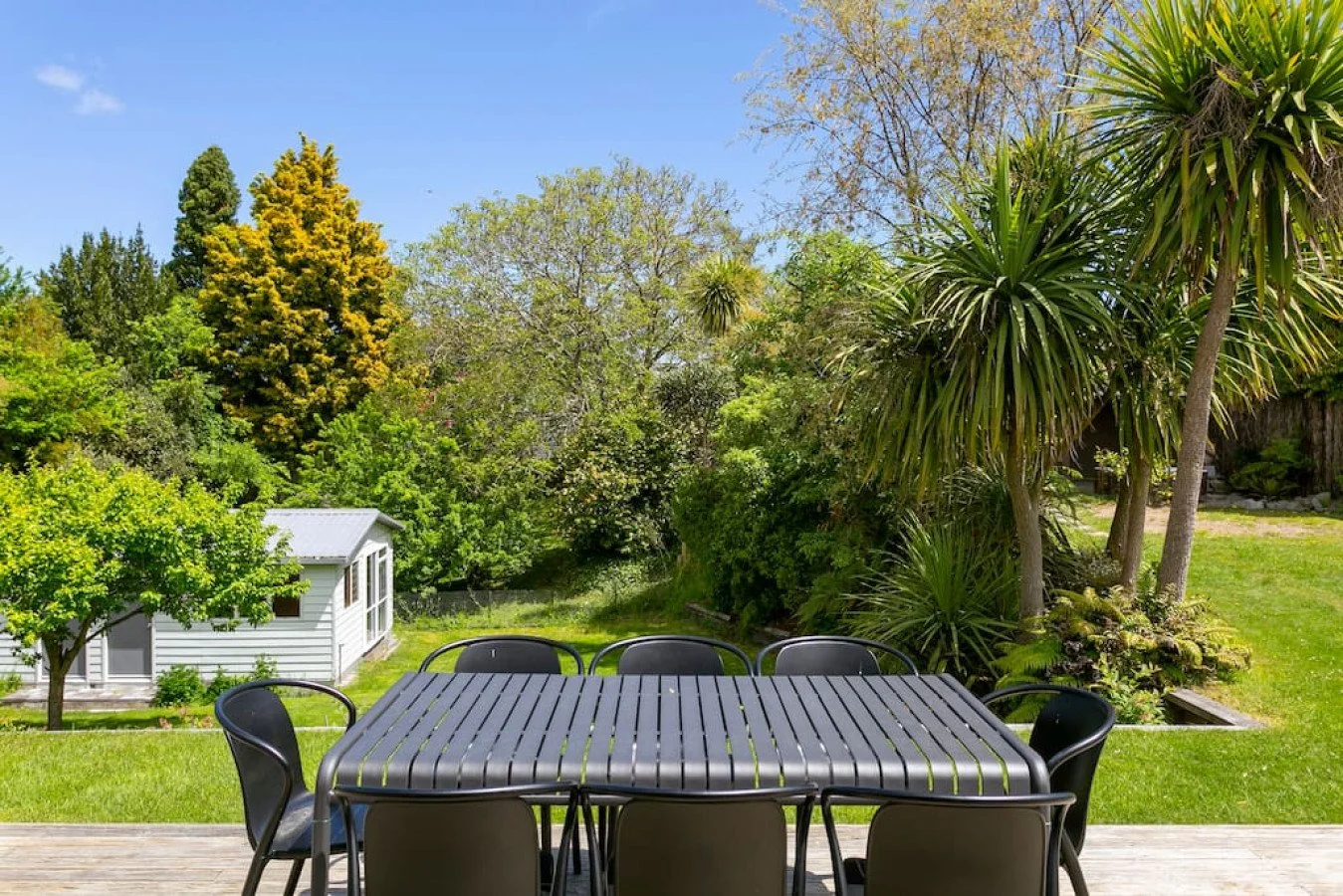 A backyard with a wooden deck, black outdoor dining table with chairs, surrounded by green grass and lush trees including palm trees and other foliage, under a clear blue sky.