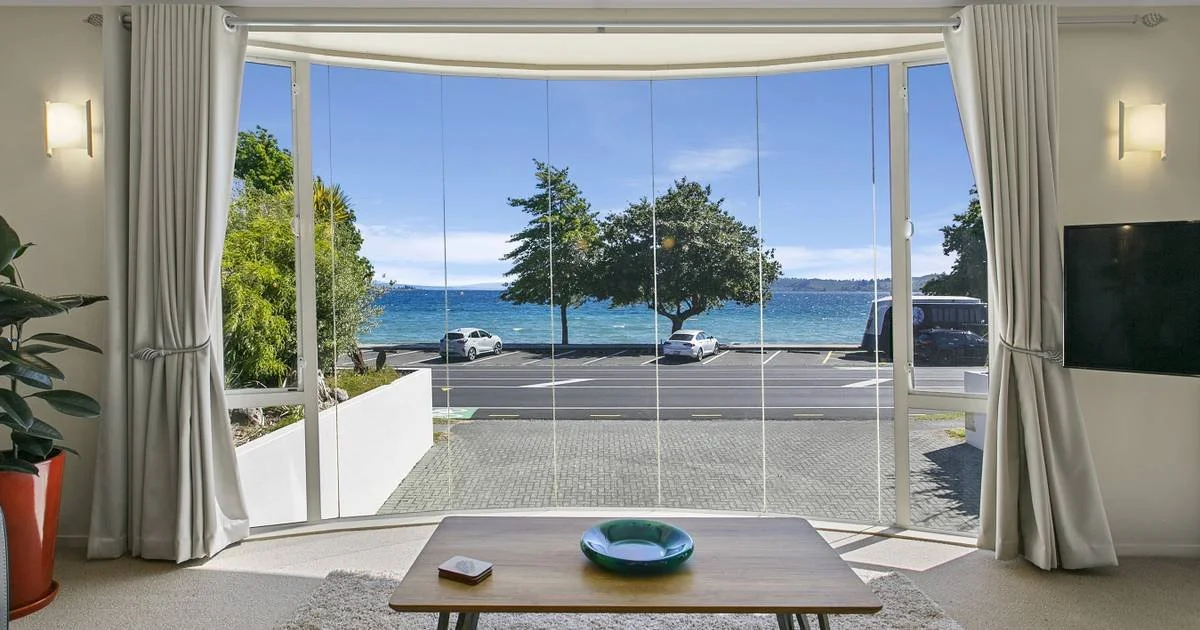 Living room with a large window showing a view of a lake, trees, parked cars, and a clear blue sky.