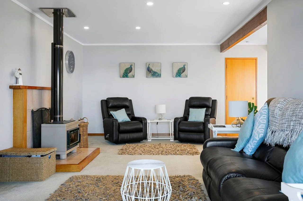 Living room with black leather recliners, a gray couch with blue pillows, a fireplace with a wood stove, a round white coffee table, and wall art of peacocks.