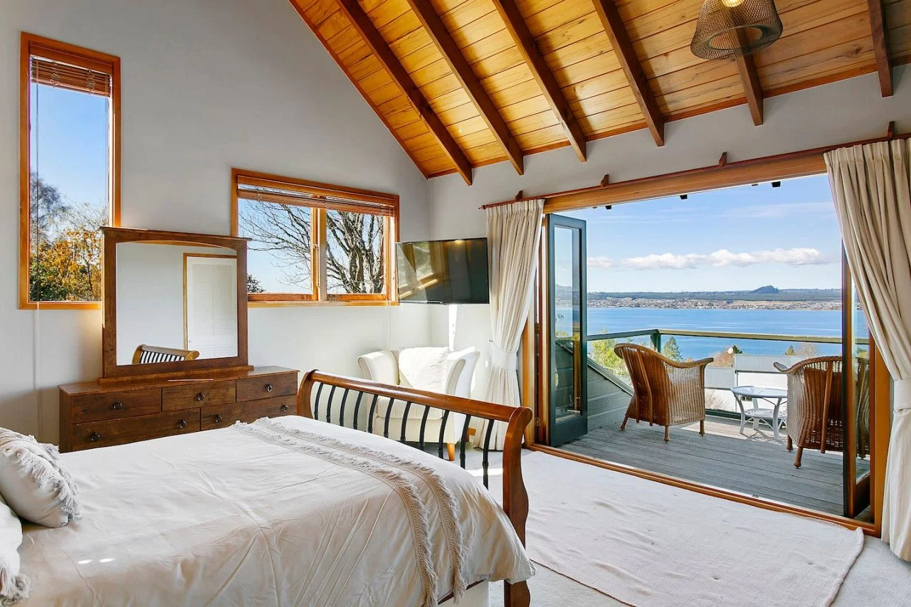 A bedroom with a wooden ceiling and large windows showing a view of water and sky. The room has a bed, dresser, mirror, and chair, with open balcony doors leading to a deck with chairs and a table.