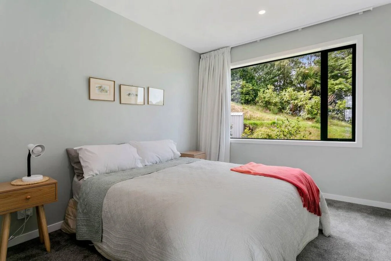 A neatly made bed in a modern bedroom with light gray walls, a wooden nightstand, a white lamp, three small framed pictures, a window showing greenery outside, and a pink blanket draped over the bed.