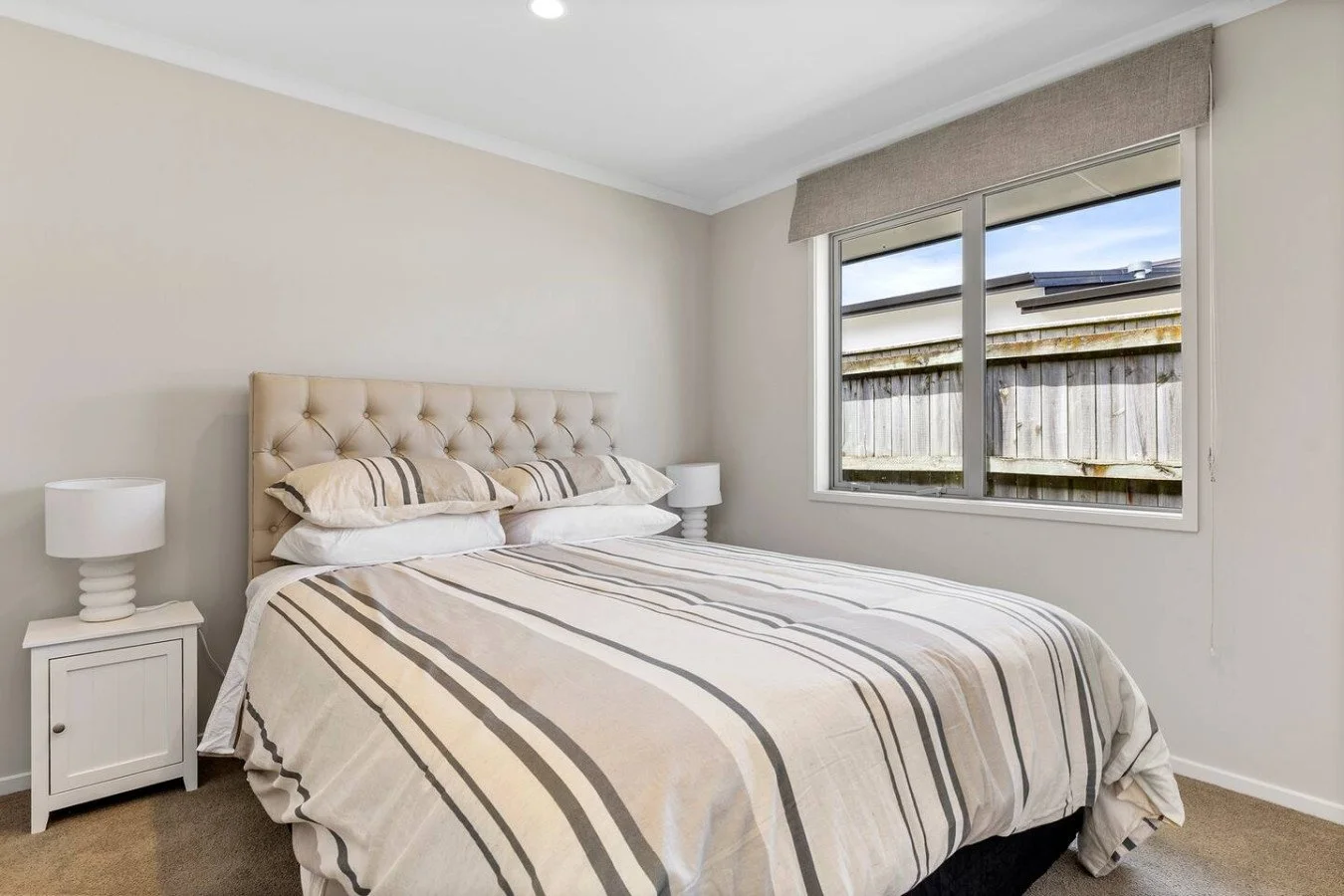A bedroom with a beige tufted headboard, a bed with striped beige, white, and brown bedding, two white nightstands with matching lamps, a window with a view of a wooden fence and a cloudy sky, beige carpet, and neutral-colored walls.