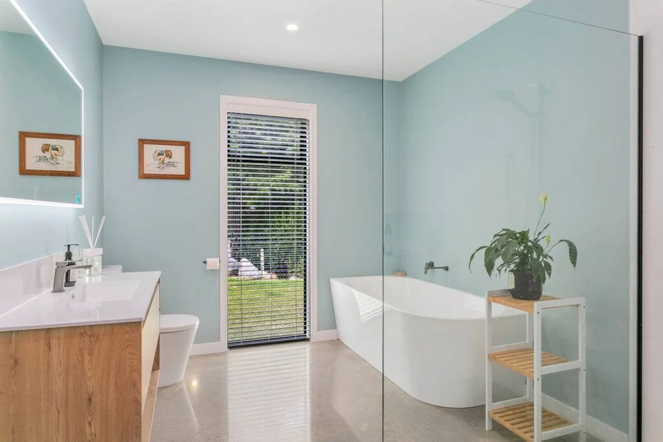 Modern bathroom with light blue walls, a bathtub, a vanity with a sink, a large mirror, a window with blinds facing outdoors, and a small wooden table with a potted plant.