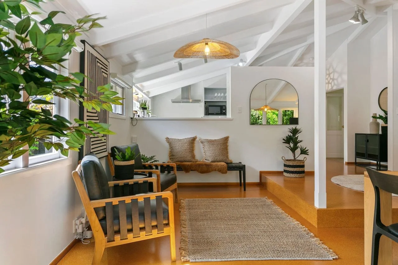 Living room with white walls, wooden floors, and a vaulted ceiling. Contains two black and wood armchairs, a small bench with beige pillows, a potted plant, a beige area rug, and decorative items.
