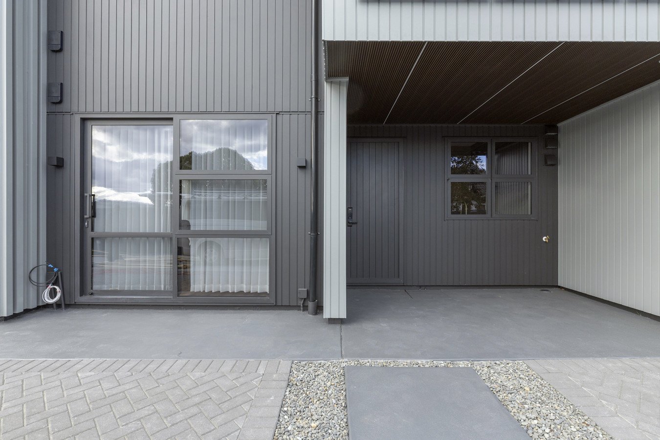 Front view of a modern building with grey metal siding, large glass doors, and windows, with a concrete and gravel walkway in front.