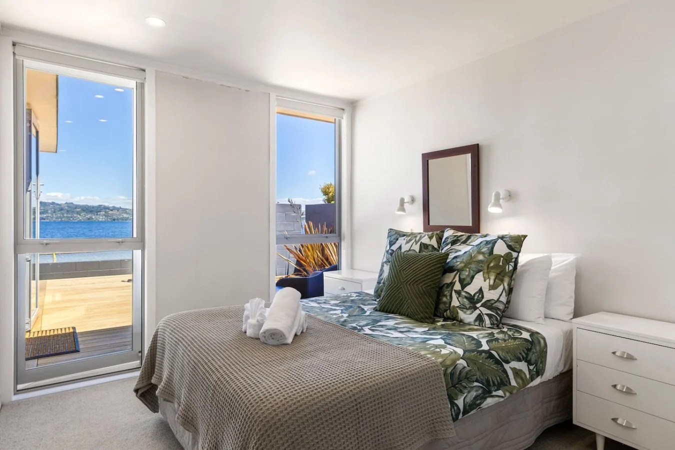 Bedroom with a bed, tropical-patterned bedding, pillows, a white nightstand, and large windows with a view of water and a deck outside.