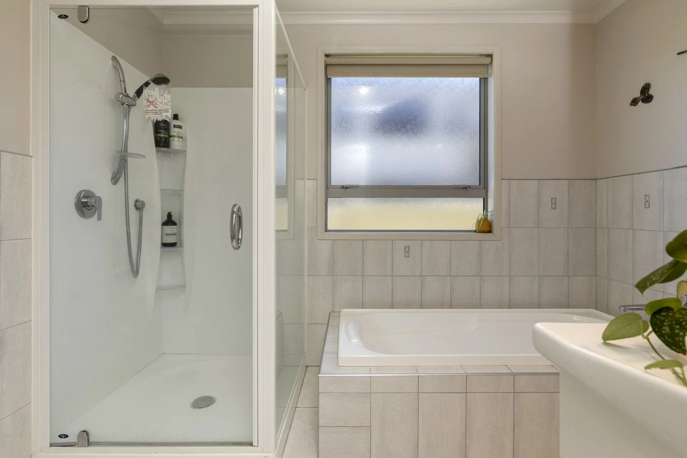 A bathroom with a walk-in shower on the left, a window with frosted glass above a bathtub in the middle, and a partial view of a sink with a plant on the right.