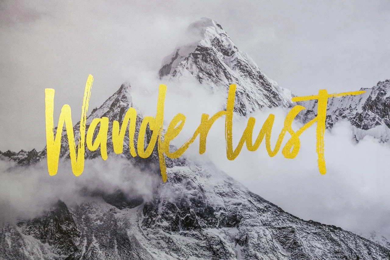 A mountain partially covered with snow and clouds, with the word "wanderlust" written in large yellow cursive text across the middle of the image.