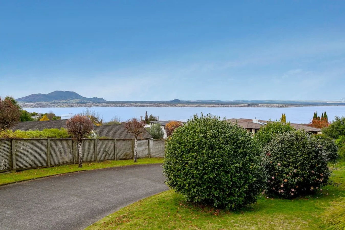 View of a residential backyard overlooking a body of water with mountains in the distance, trees, and shrubs in the foreground, and a fence separating the yard from neighboring houses.