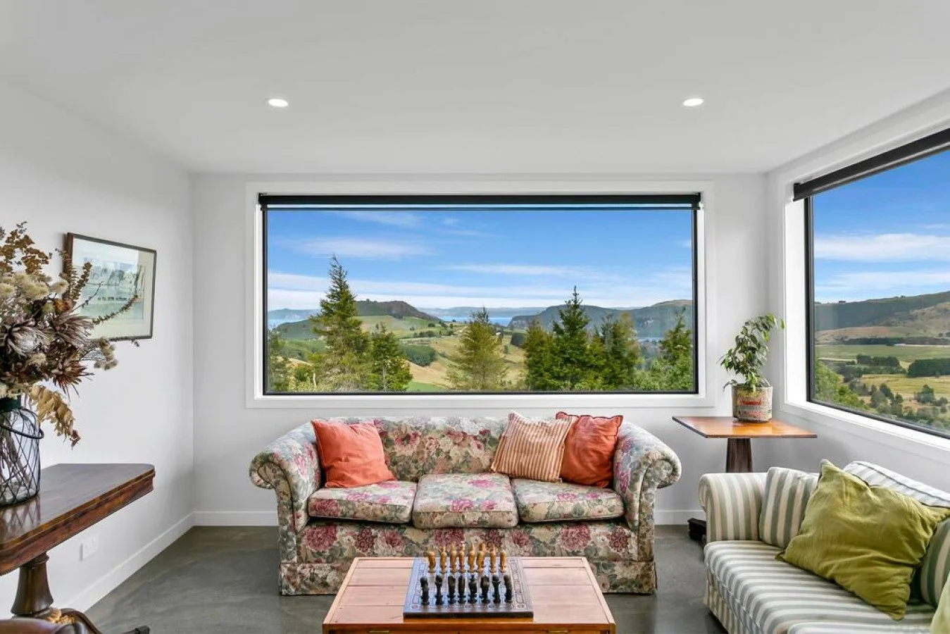 Living room with floral sofa, striped armchair, wooden coffee table with chessboard, large windows showing scenic hills and trees, potted plant, framed picture on left wall, and various cushions.