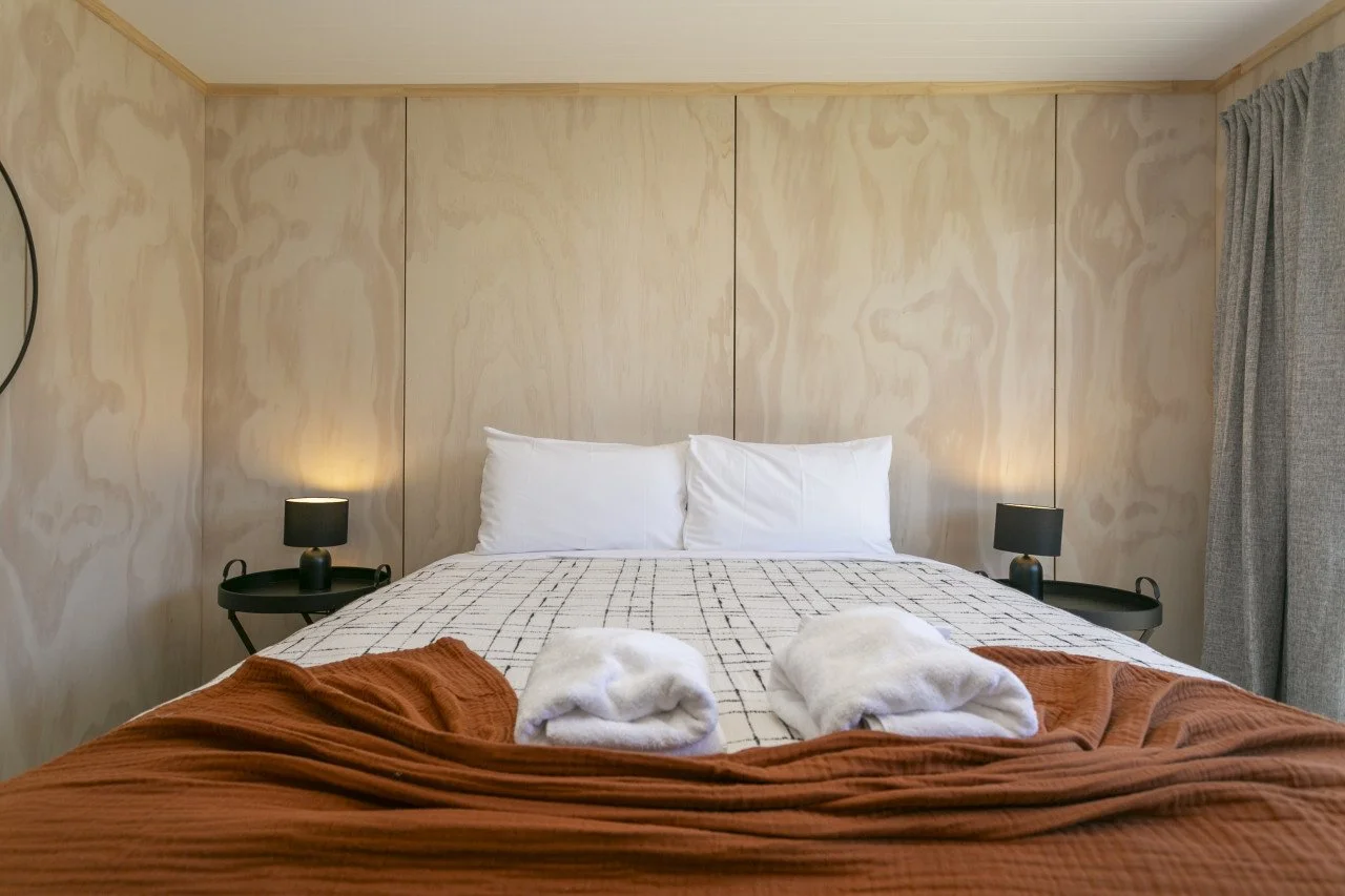 Hotel bed with white pillows, folded white towels, checkered bedsheet, rust-colored blanket, two black bedside lamps, wood-paneled wall, gray curtains, and circular wall decor.
