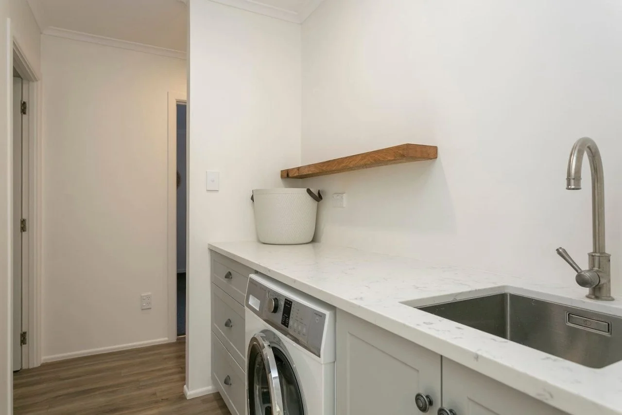 Laundry room with white cabinetry, marble countertop, stainless steel sink, and washing machine. A wooden shelf and a basket are on the counter, with a doorway in the background.