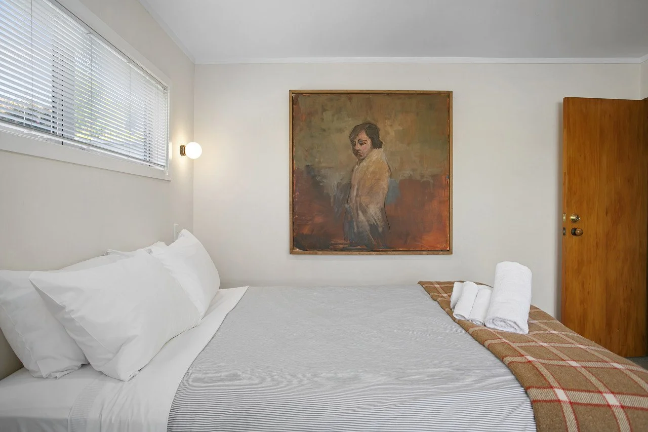 A minimalist hotel room with a bed covered in white linens, accented by folded towels and a blanket. There is a window with blinds, a wall-mounted light, and a large framed painting of a person on the wall.