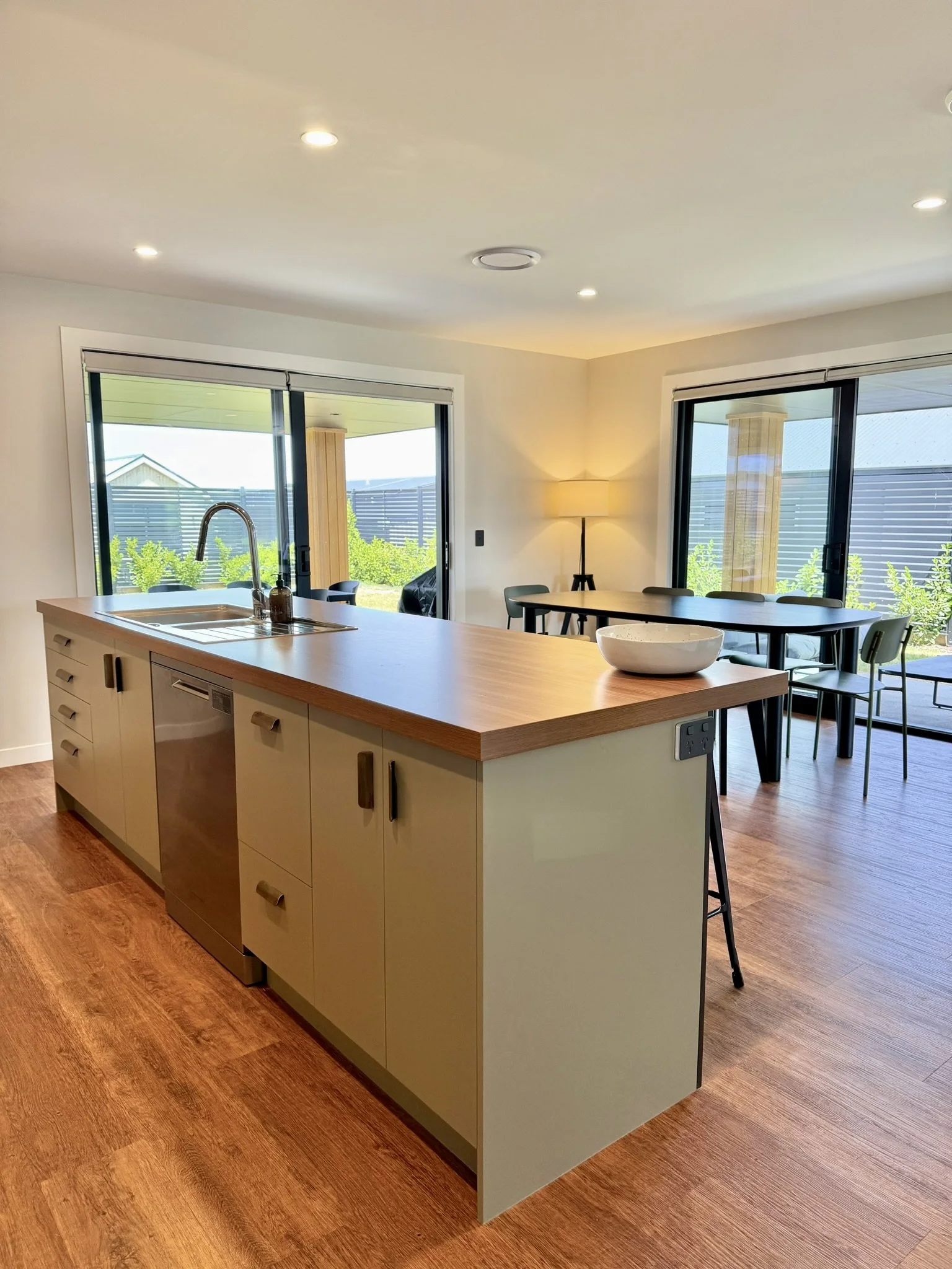 Modern kitchen with an island, wooden countertop, and a sink, adjacent to a dining area with large windows and sliding glass doors, hardwood flooring, and a sunlit outdoor patio.