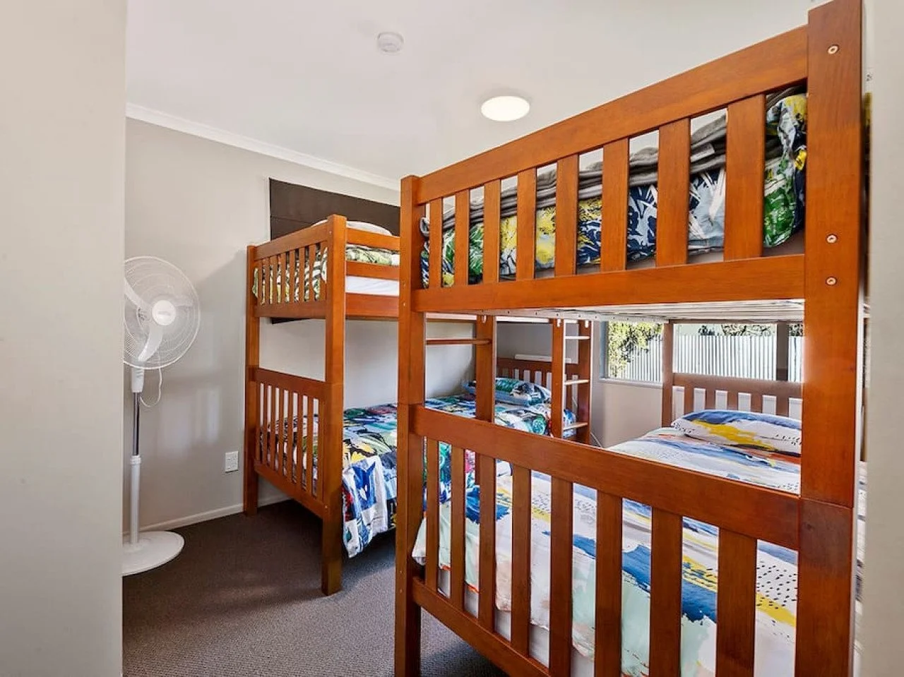 A bedroom with two wooden bunk beds, each with a twin mattress and colorful bedding. A white standing fan is in the corner on the left, and windows with blinds are at the back.