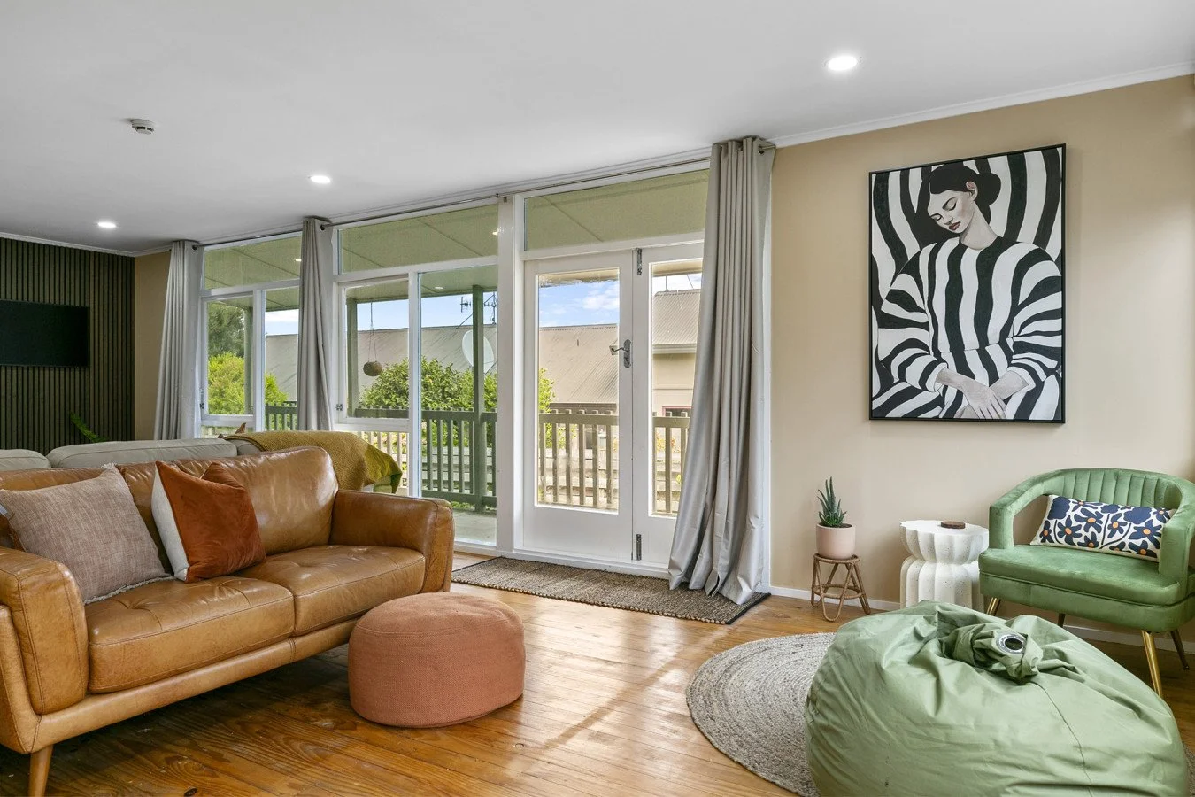 Living room with leather sofa, green armchair, round rug, large windows with curtains, wall art of a woman in black and white stripes, and wood flooring.