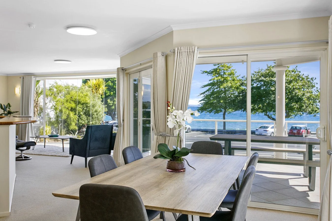 Living and dining room with large glass doors and windows overlooking a lake with parked cars and trees