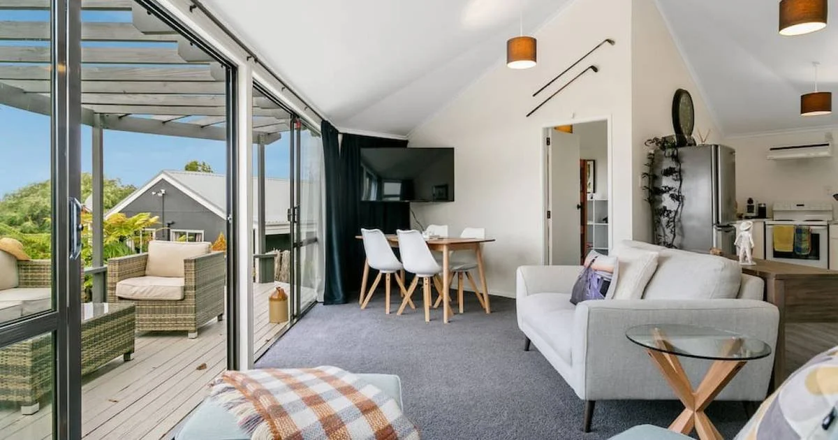 A living room with a large glass sliding door leading to an outdoor patio with wicker chairs. Inside, there is a white sofa, a glass side table, a dining table with four chairs, a television, and kitchen area in the background. The room has white wal