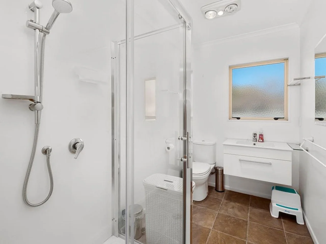 A clean, modern bathroom with a shower stall, toilet, white vanity with mirror, frosted windows, and brown tiled floor.