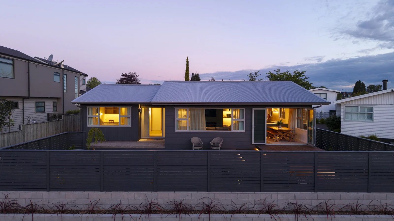 A modern house at dusk with warm yellow interior lights visible through the windows and sliding doors, surrounded by a black fence and a garden in the foreground.