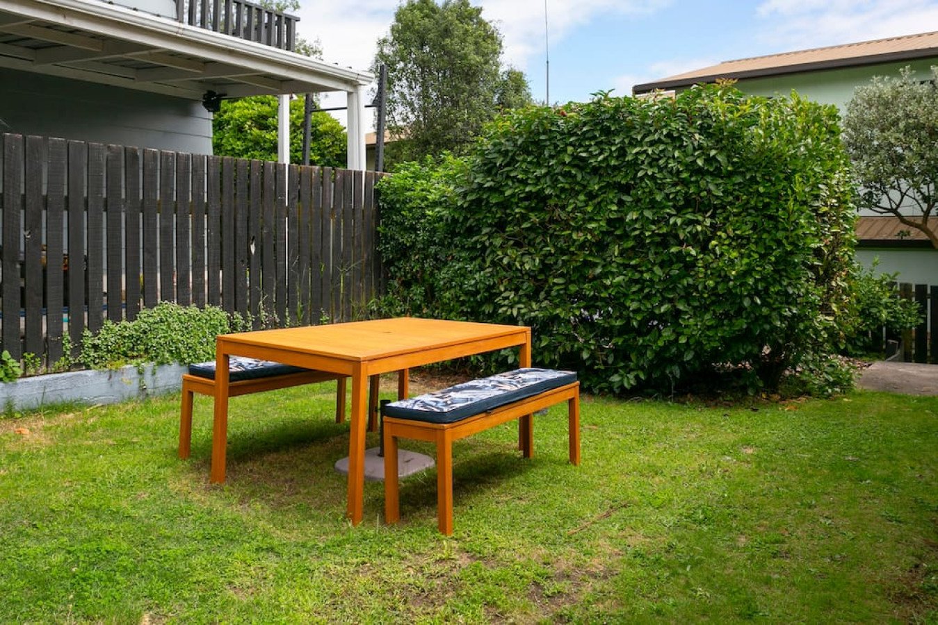 Small backyard with a wooden table and two matching benches with cushions on a grassy lawn, surrounded by bushes and a wooden fence.