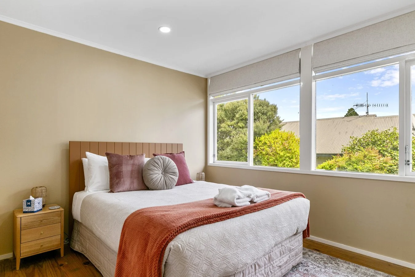 Bedroom with a bed, pillows, and large windows showing trees and a roof outside.