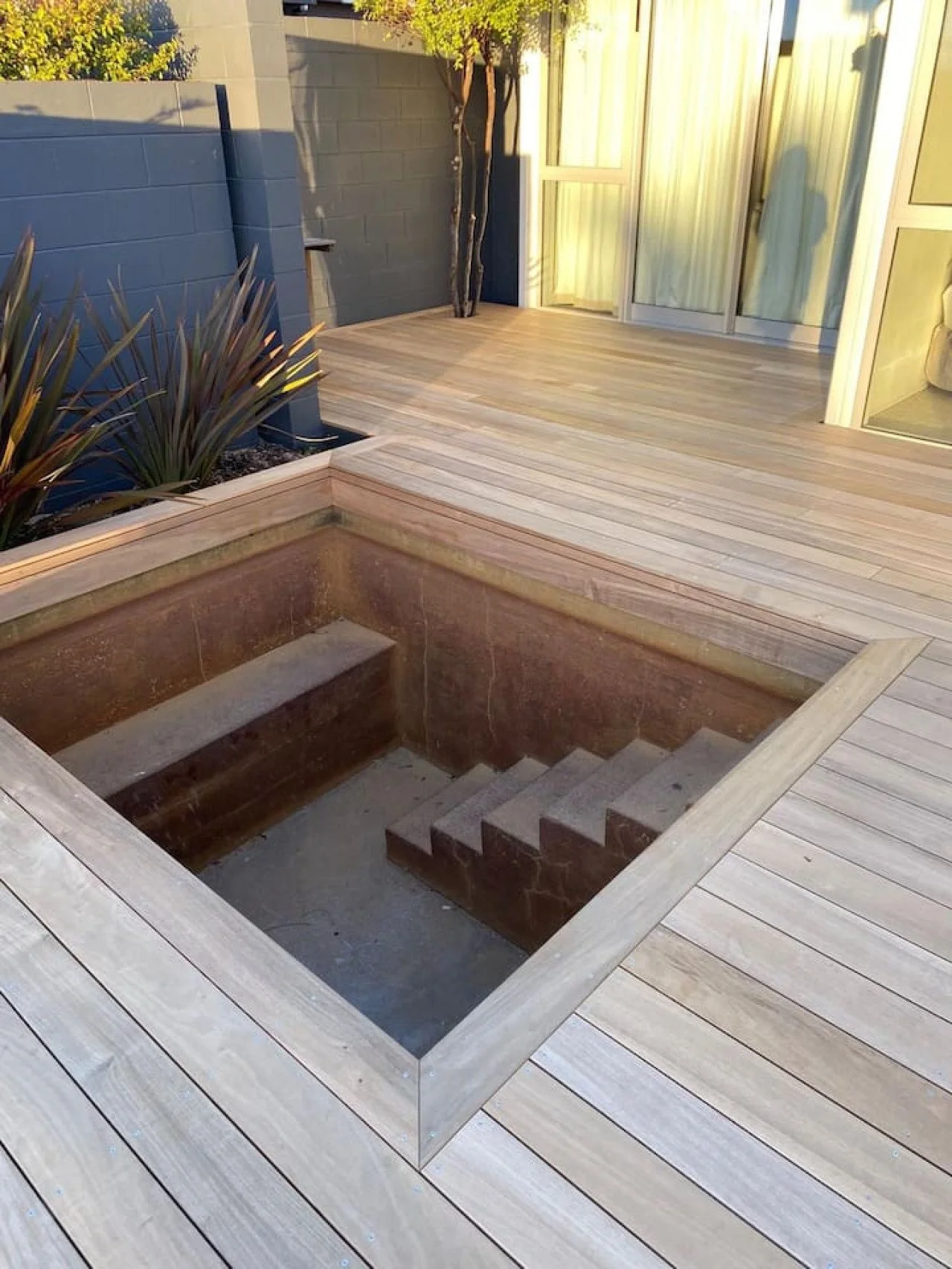 An outdoor wooden deck with a built-in, empty, sunken step or hot tub with brick interior and stairs, next to a plant with spiky leaves, enclosed by a gray wall and glass sliding doors.