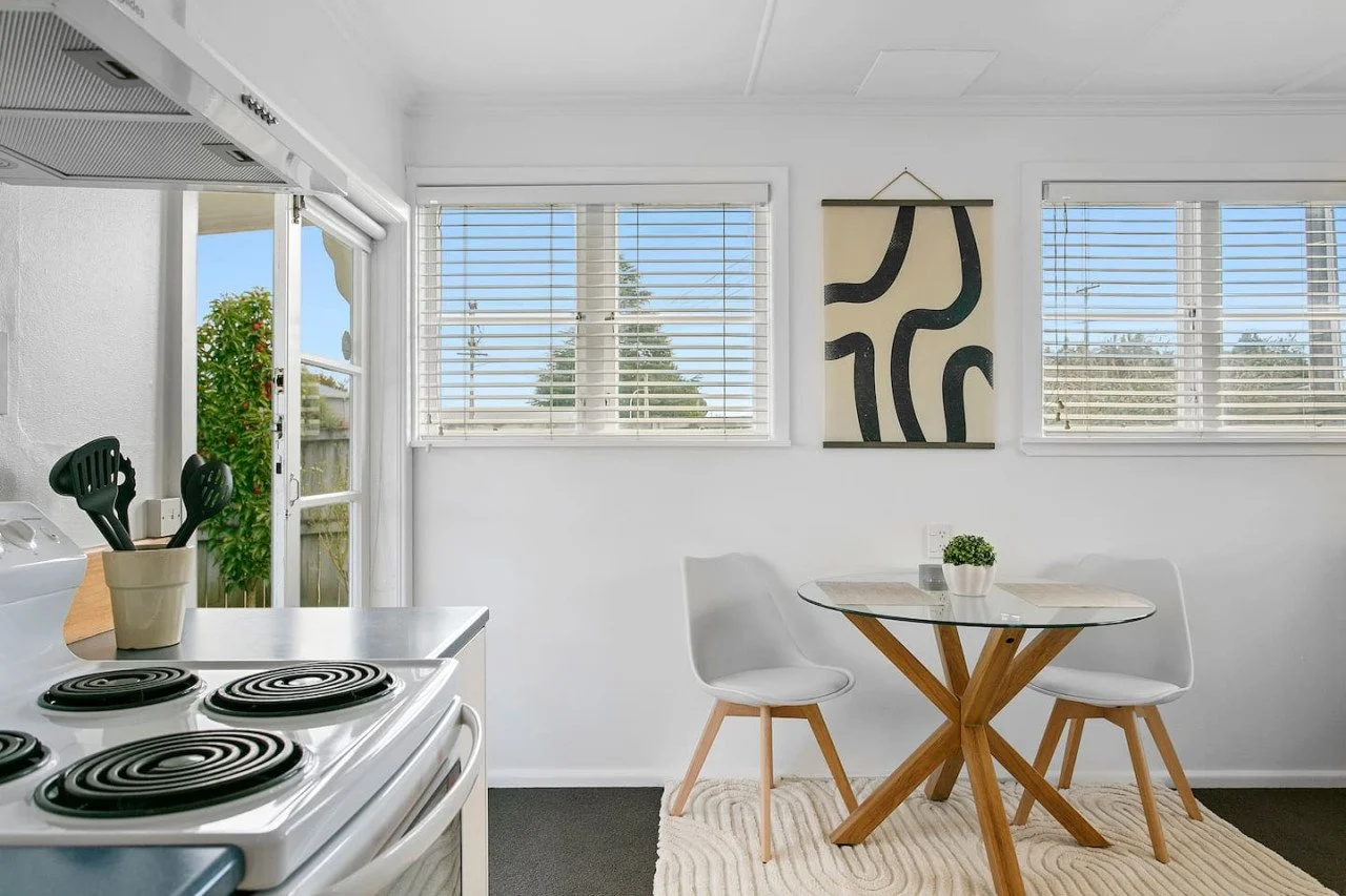 A bright, modern kitchen and dining area with white walls and windows with blinds, a glass-top round table with wooden legs, and two white chairs. There's a small potted plant on the table, a piece of abstract wall art, and a kitchen counter with coo