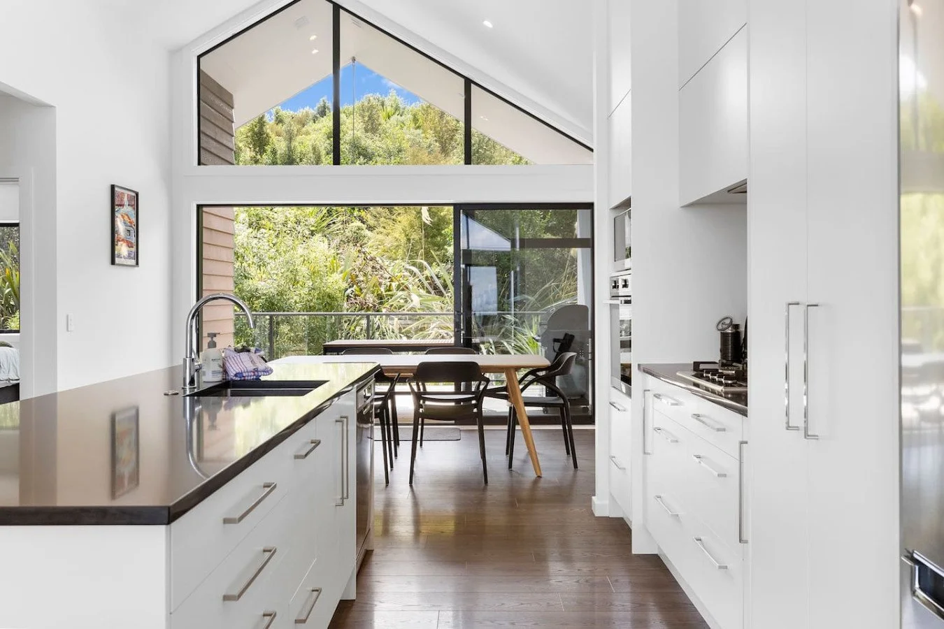 Modern kitchen with large windows overlooking greenery, white cabinets, a black countertop on an island with a sink, and a dining area with a wooden table and black chairs in the background.