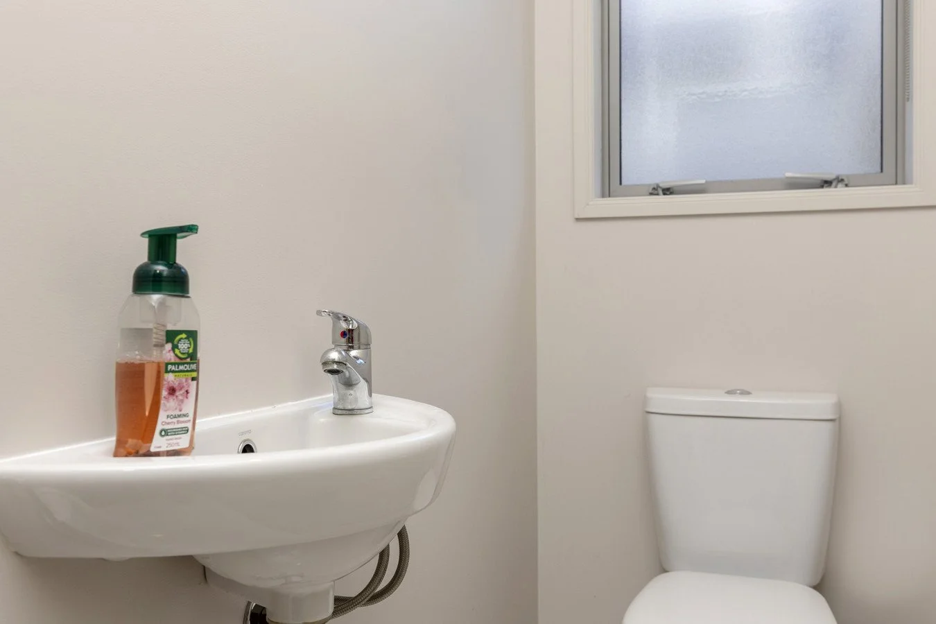A small white bathroom with a wall-mounted sink, a chrome faucet, a soap dispenser, a toilet, and a frosted window.