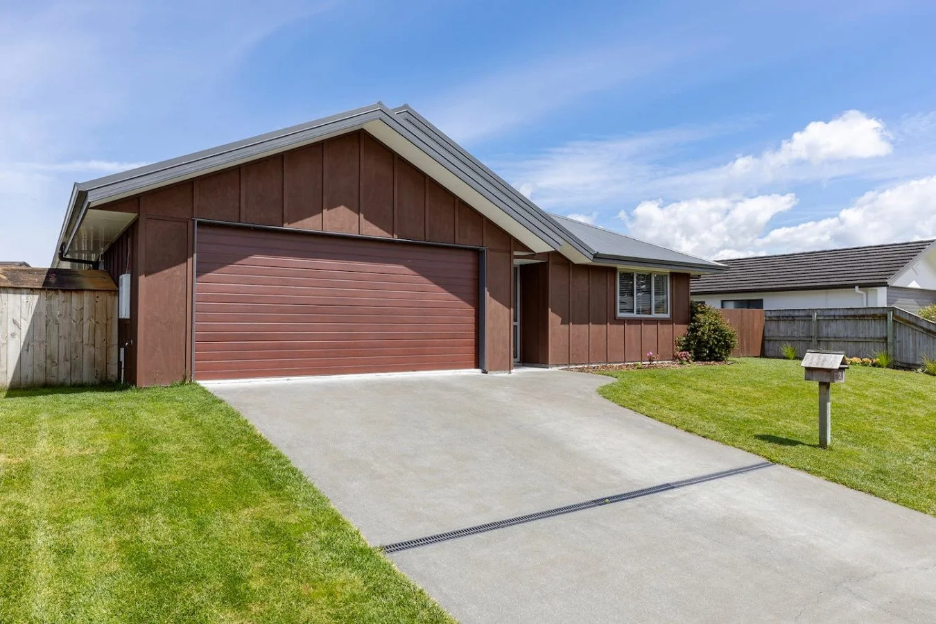 Front view of a modern house with a brown garage door, wooden siding, a driveway, and a front lawn, with neighboring houses in the background and a blue sky with clouds.