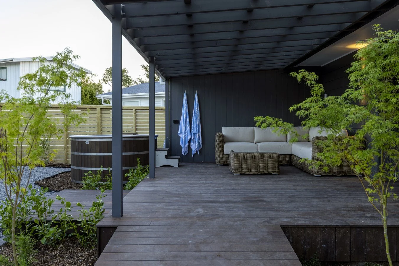 Outdoor patio with wicker seating, blue and white striped towels hanging, trees, and a hot tub near a wooden fence.